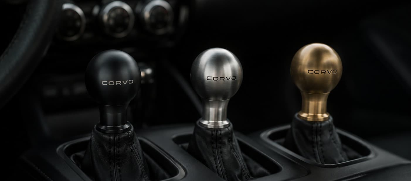 Three CORVO-001 shift knobs in black anodized aluminum, raw titanium, and brass, mounted in a dark car interior. Concept render.